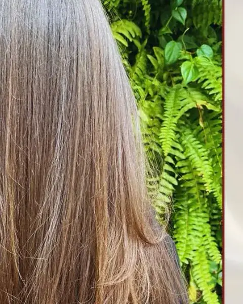 Cabelo longo com coloração e reflexos claros, parede verde de plantas ao fundo