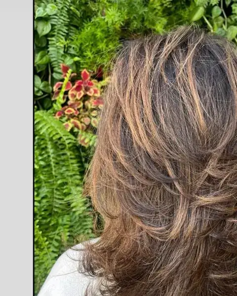 Cabelo com escova e luzes caramelo, parede verde de plantas ao fundo
