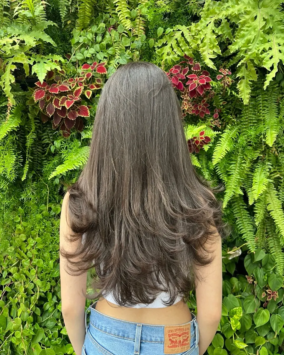 Cabelo longo com mechas claras, cliente de costas, parede verde de plantas ao fundo